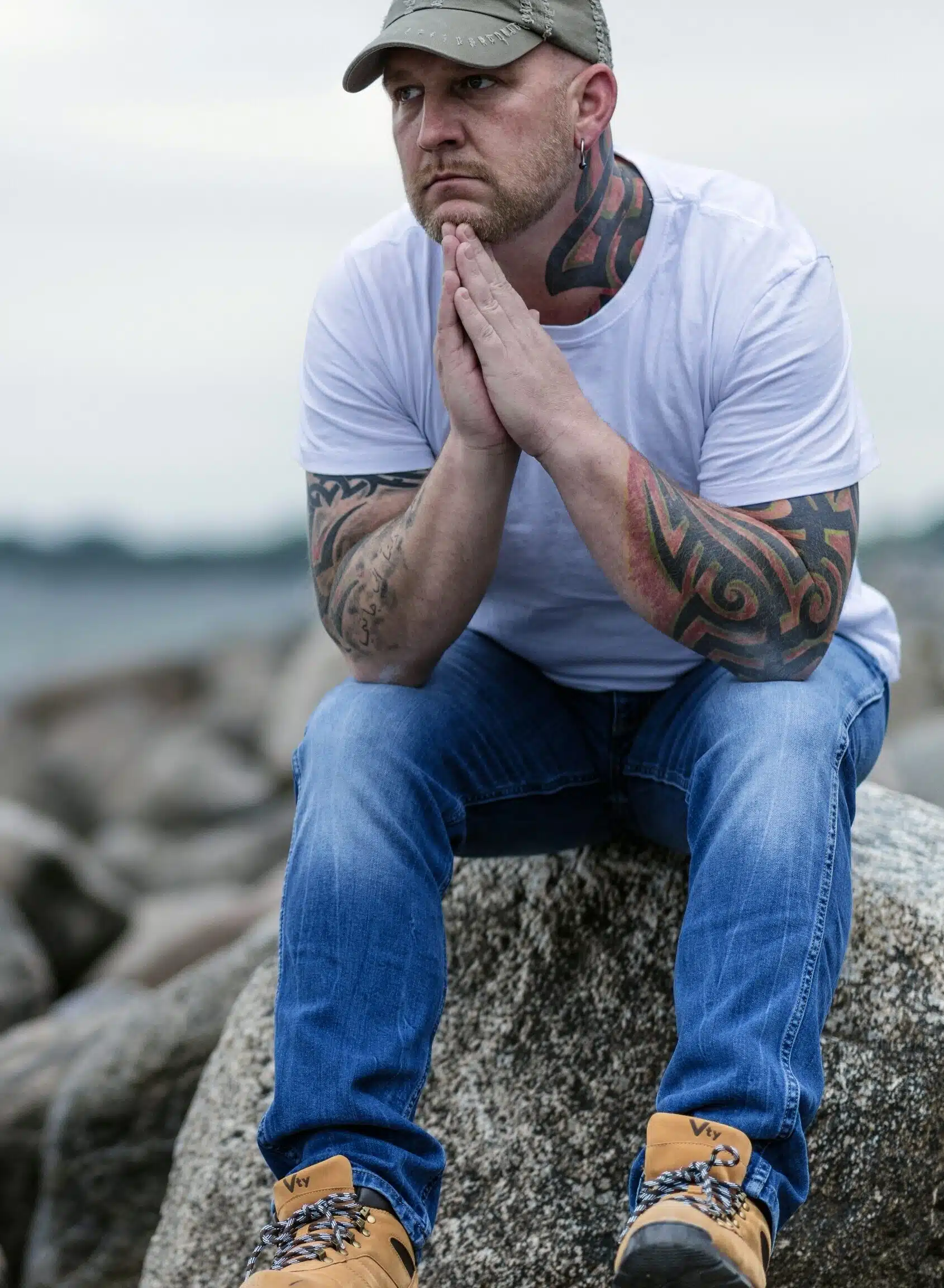 Post's photo of man with tattoos sitting on a rock by the water in prayer type of pose with eyes open thinking about online borderline treatment options. Solemn, flat facial expression.